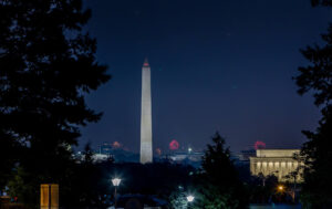 Professional Photographer The Washington Monument stands illuminated at night, framed by trees. Fireworks burst in the sky behind it, captured by a photographer, and the Lincoln Memorial is visible to the right, also lit up against the dark blue sky. www.maranonimagery.com
