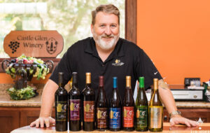 Professional Photographer A man with a beard, wearing a black Castle Glen Winery shirt, stands behind a counter displaying eight bottles of wine. A Castle Glen Winery sign is on the wall behind him—perfect for headshots or capturing special moments like wedding photography. www.maranonimagery.com
