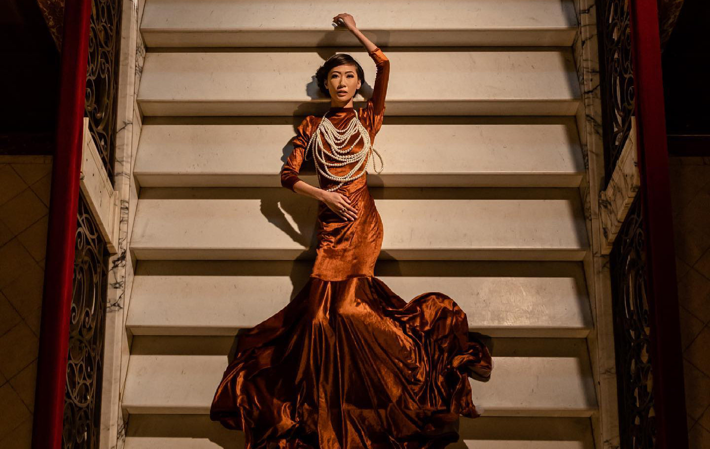 Professional Photographer A woman in a dramatic, shiny brown gown with a long train and layered pearl necklaces lies elegantly across white stairs, one arm raised above her head—captured by a skilled photographer—framed by ornate railings. www.maranonimagery.com