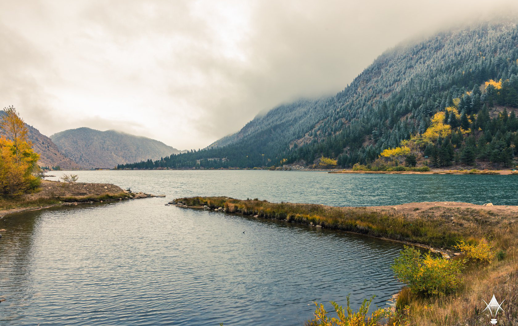 Professional Photographer A serene lake bordered by pine-covered mountains with light snow and autumn foliage, under a cloudy sky. The gently curving shoreline offers a stunning backdrop, ideal for a photographer seeking inspiration or unique wedding photography. www.maranonimagery.com