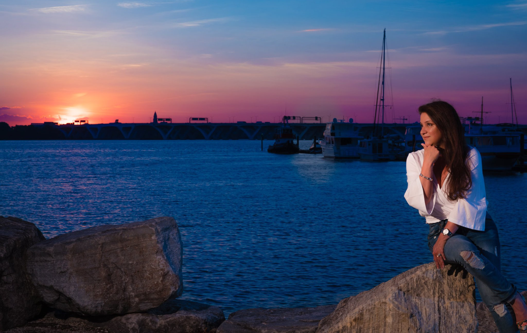 Professional Photographer A woman in a white shirt and jeans sits on large rocks by the water at sunset, captured beautifully by a photographer. Boats and a distant bridge are visible in the background under a colorful sky. www.maranonimagery.com