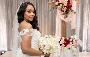Professional Photographer A bride in an off-shoulder lace dress holds a white bouquet and stands beside a flower-adorned wooden cross, looking over her shoulder and smiling softly—a charming moment captured in elegant wedding photography. www.maranonimagery.com