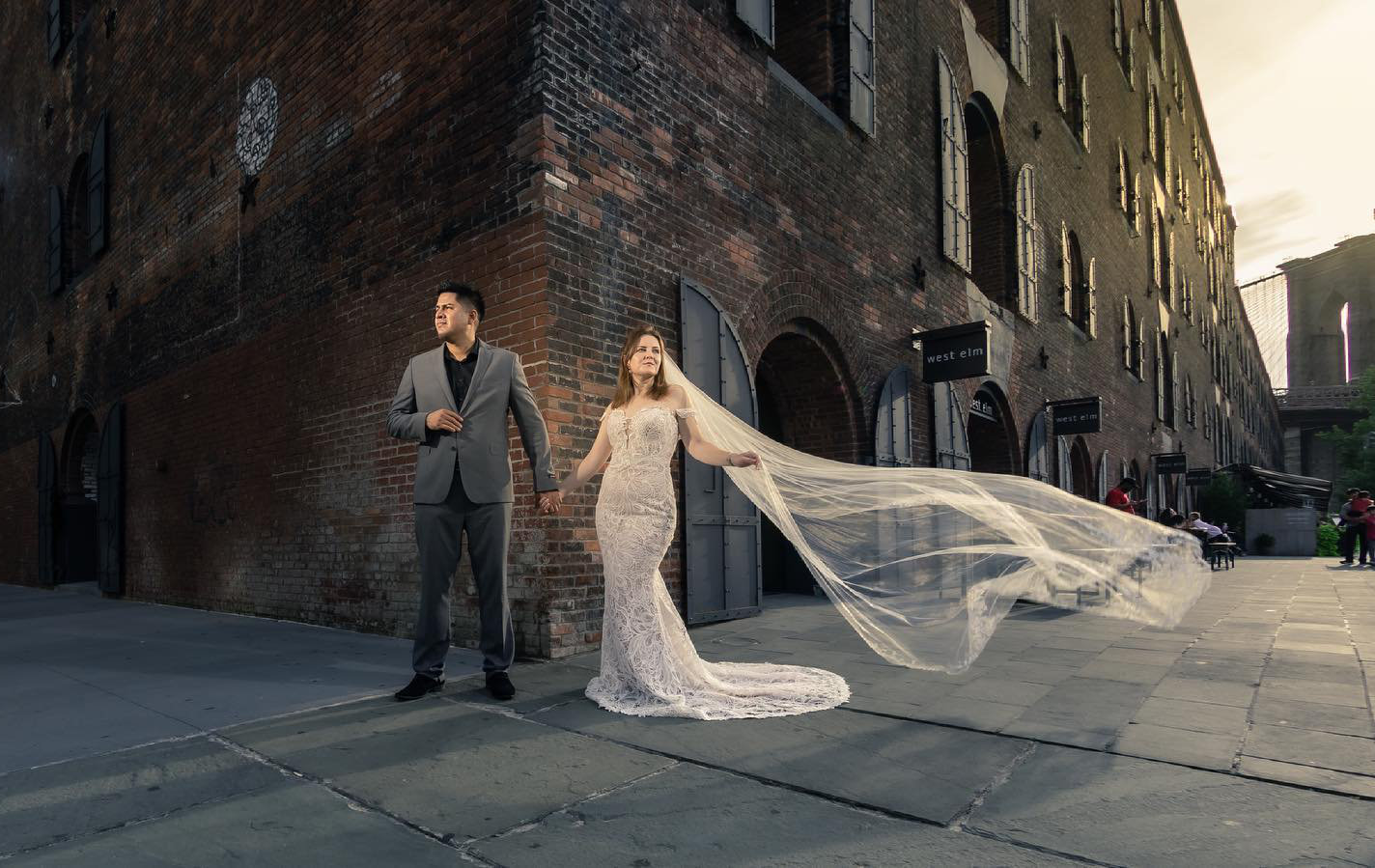 Professional Photographer A bride in a lace gown with a long, flowing veil holds hands with a groom in a suit, standing outside a brick building at sunset—perfectly captured by skilled wedding photography as soft light illuminates their moment. www.maranonimagery.com