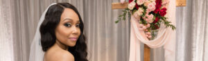 Professional Photographer A bride in a white dress and veil stands before a wooden cross adorned with pink and red flowers, looking over her shoulder and smiling softly. Perfect for Wedding Photography, silver curtains shimmer gently in the background. www.maranonimagery.com