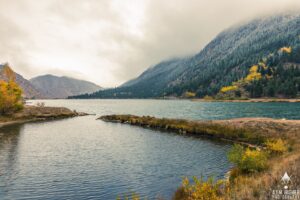 Professional Photographer A serene lake surrounded by forested mountains with autumn foliage under a cloudy sky. Mist partially covers the mountain peaks, and a photographer captures the calm water reflecting the tranquil atmosphere. www.maranonimagery.com