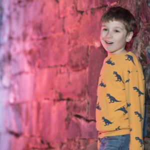 Professional Photographer A smiling child with short hair poses for headshots, wearing a yellow sweatshirt with dinosaur prints, standing against a stone wall illuminated by pink and purple lighting. www.maranonimagery.com