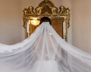 Professional Photographer A bride stands facing a large ornate mirror, her back to the camera, wearing a white dress and flowing veil—an elegant moment beautifully captured by a wedding photographer in a softly lit room. www.maranonimagery.com