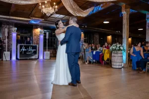 Professional Photographer A bride and groom share their first dance in a warmly lit venue with wood floors, exposed brick, and guests seated at tables. A photographer captures the moment as a DJ plays in the background beneath draped fabric and a chandelier. www.maranonimagery.com