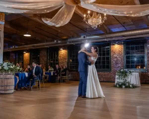 Professional Photographer A bride and groom share their first dance in a rustic, warmly lit venue while a photographer captures candid portraits. Exposed brick walls, wooden beams, and draped white fabric create a romantic setting as guests watch from flower-adorned tables. www.maranonimagery.com