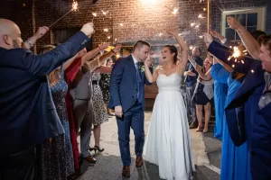 Professional Photographer A bride and groom walk hand in hand, smiling and celebrating, as guests on both sides hold sparklers to create a festive tunnel—capturing the joy of this magical moment in stunning wedding photography. www.maranonimagery.com