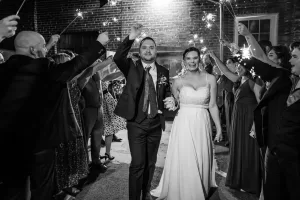 Professional Photographer A bride and groom walk hand in hand through a crowd of guests holding sparklers, smiling joyfully. Captured by a talented photographer, the black and white scene radiates celebration for the couple’s wedding. www.maranonimagery.com