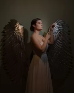 Professional Photographer A woman in a white dress stands in front of a dark background, posing with her arms raised. Large, metallic, silver angel wings are attached to the wall behind her, creating the illusion she has wings—perfect for creative wedding photography. www.maranonimagery.com