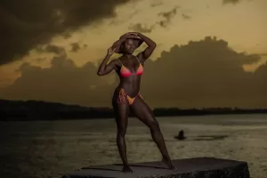 Professional Photographer A woman in a pink bikini and wide-brim hat poses confidently on a concrete platform by the water at sunset, as a photographer captures stunning portraits with dark clouds and distant land visible in the background. www.maranonimagery.com