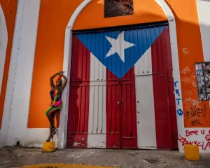 Professional Photographer A woman in a colorful dress poses beside a large door painted with the Puerto Rican flag, set in an orange and white building covered with graffiti and stickers—perfect inspiration for any photographer seeking unique city scenes. www.maranonimagery.com