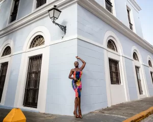 Professional Photographer A woman in a colorful, fitted dress stands confidently at the corner of a pale blue building with arched windows and a lamp above her, posing with one arm raised for striking portraits. www.maranonimagery.com