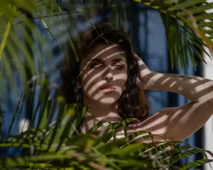 Professional Photographer A woman stands behind green palm leaves, with sunlight casting leaf-shaped shadows across her face and body. Perfect for creative portraits, she gazes ahead with one hand behind her head against a striking blue background. www.maranonimagery.com