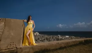 Professional Photographer A woman in a flowing yellow dress poses against a stone wall, as a photographer captures her overlooking a cemetery by the ocean under a clear, blue sky. www.maranonimagery.com