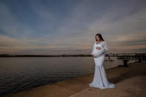 Professional Photographer A photographer captures a pregnant woman in a long white dress by the waterfront at sunset, gently holding her belly and smiling. A bridge and birds are visible in the background beneath a cloudy sky. www.maranonimagery.com