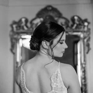 Professional Photographer A woman in a lace dress stands with her back to the camera, looking over her shoulder. Her dark hair is styled in an elegant updo. Captured by a talented photographer, she poses before a large, ornate mirror in this black and white image. www.maranonimagery.com
