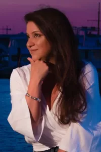 Professional Photographer A woman with long brown hair, wearing a white blouse and a silver bracelet, sits by the water at sunset with her hand resting on her chin, looking thoughtfully to the side—like she’s posing for a photographer specializing in headshots. www.maranonimagery.com