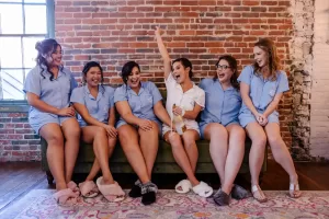 Professional Photographer Six women in matching blue pajamas and slippers sit on a couch in front of a brick wall, laughing. The woman in the center wears white, holding up a champagne bottle—capturing a joyful moment of wedding photography at its finest. www.maranonimagery.com