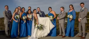 Professional Photographer A bride in a white gown is held up by the groom and surrounded by a smiling wedding party. Captured by a talented wedding photographer, the bridesmaids in blue dresses and groomsmen in gray suits stand outdoors on a sunny day, holding bouquets. www.maranonimagery.com