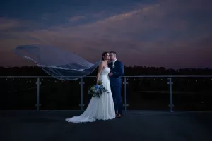 Professional Photographer A bride and groom stand on a balcony at dusk. The bride’s veil flows in the wind as they embrace tenderly, framed by a dark treeline and pink-purple sky. A photographer captures this romantic portrait as the bride holds her bouquet of flowers. www.maranonimagery.com