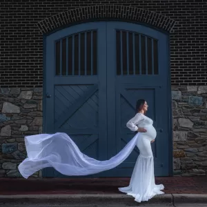 Professional Photographer A pregnant woman in a long, flowing white dress stands sideways in front of a large blue door and stone wall, the train of her dress billowing behind her in this stunning portrait by a talented wedding photographer. www.maranonimagery.com