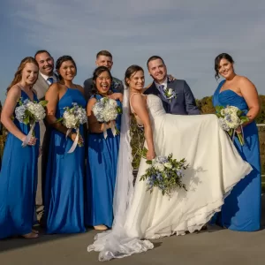 Professional Photographer A bride in a white dress is being held up by the groom, surrounded by smiling bridesmaids in blue dresses and groomsmen, all holding bouquets and posing outdoors on a sunny day for joyful portraits. www.maranonimagery.com