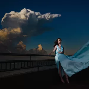 Professional Photographer A photographer captures a woman in a flowing light blue dress standing on a balcony at dusk, dramatic clouds behind her. The wind stirs her dress and hair, infusing the portrait with movement and elegance. www.maranonimagery.com