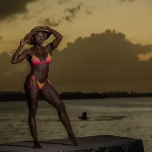 Professional Photographer A woman in a colorful bikini stands on a concrete platform by the water at sunset, holding a wide-brimmed hat, while a photographer captures the dramatic clouds and boat in the background. www.maranonimagery.com