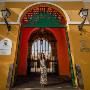 Professional Photographer A woman in a floral outfit stands in front of a large, colorful arched entrance—perfect for creative portraits—with yellow walls, a red and green archway, black gates, and Museo de San Juan signs on either side. www.maranonimagery.com