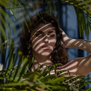 Professional Photographer A woman with long dark hair stands among palm leaves. Sunlight casts striped shadows across her face and body, creating a striking headshot. She gazes ahead with one hand touching her hair, the background softly out of focus. www.maranonimagery.com