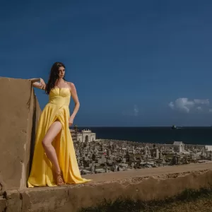Professional Photographer A woman in a bright yellow dress stands against a stone wall, overlooking a large cemetery by the sea under a sunny blue sky—a picturesque moment perfect for creative portraits by an experienced photographer. www.maranonimagery.com