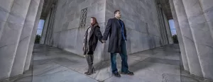 Professional Photographer A woman and a man stand back to back, holding hands, in front of a large stone building with columns. Captured by a skilled photographer, both look away from each other in coats and boots, on a cloudy day. www.maranonimagery.com