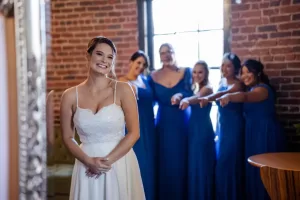 Professional Photographer A bride in a white dress smiles in the foreground while five bridesmaids in blue dresses stand behind her, smiling and pointing excitedly toward her—captured perfectly by a talented photographer in a room with brick walls and a large window. www.maranonimagery.com