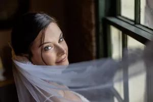 Professional Photographer A bride with dark hair and bright eyes looks over her shoulder, smiling gently, as her veil flows in soft light—beautifully captured by a wedding photography expert beside a sunlit window. www.maranonimagery.com