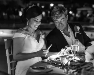 Professional Photographer A bride in a white dress and a man in a suit sit together at a table, smiling and looking at the phone in the bride's hand; the scene is warmly lit, capturing a joyful wedding photography moment with glasses and plates on the table. www.maranonimagery.com