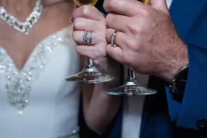 Professional Photographer A bride and groom, wearing wedding bands and elegant jewelry, hold wine glasses together in formal attire as a photographer captures their joyful celebration. www.maranonimagery.com