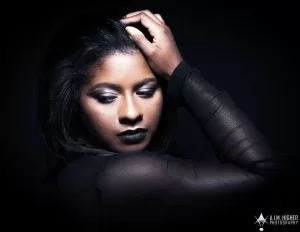 Professional Photographer A woman with dark hair and dramatic makeup poses with one hand on her head against a black background, wearing a sheer black top and looking down with eyes closed—an evocative portrait by a skilled photographer. www.maranonimagery.com