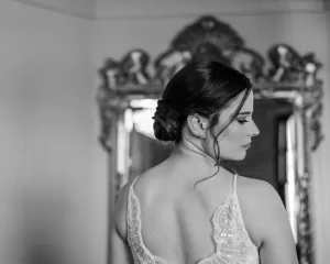 Professional Photographer A woman in a lace-trimmed dress stands with her back to the camera, glancing to the side. Her dark hair is styled in an elegant updo, evoking timeless wedding photography. An ornate mirror graces the black and white scene. www.maranonimagery.com