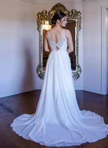 Professional Photographer A woman in a flowing white wedding dress with a lace back stands facing an ornate mirror on a wooden floor in a light-colored room, perfect for elegant wedding photography or timeless bridal portraits. www.maranonimagery.com