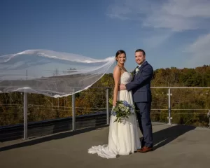 Professional Photographer A bride and groom stand on an outdoor balcony with trees in the background. Their smiles capture the joy of wedding photography as the bride's veil blows in the wind, holding a bouquet under a clear blue sky. www.maranonimagery.com