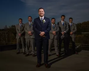 Professional Photographer A groom in a navy suit and boutonniere stands smiling in front of five groomsmen in gray suits with blue ties, all posing outdoors under a partly cloudy sky—an elegant moment captured through wedding photography. www.maranonimagery.com