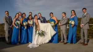 Professional Photographer A wedding photography session captures a joyful rooftop party. The bride and groom are in the center, with the groom lifting the bride. Bridesmaids in blue dresses and groomsmen in gray suits with blue ties all smile at the camera. www.maranonimagery.com