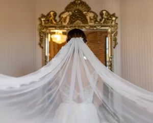 Professional Photographer A bride in a white dress with a long, flowing veil stands facing an ornate gold mirror, her back to the camera, in a softly lit room—capturing the timeless elegance often sought in portraits and headshots. www.maranonimagery.com