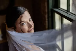 Professional Photographer A bride with dark hair and blue eyes looks over her shoulder and smiles softly, her white veil flowing as she stands by a window with green frames—an enchanting moment captured by the photographer in stunning portraits. www.maranonimagery.com