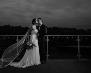 Professional Photographer A bride and groom stand close together on an outdoor balcony at dusk. Captured by a skilled photographer, the groom kisses the bride’s cheek while she holds a bouquet and her veil flows behind her in this black and white scene. www.maranonimagery.com