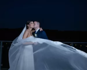Professional Photographer A photographer captures a bride and groom embracing at night on a balcony. The groom kisses her cheek as her veil flows dramatically in the wind, creating romantic portraits against a dark, outdoor background. www.maranonimagery.com