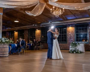 Professional Photographer A bride and groom share their first dance in a rustic, warmly lit venue with wooden beams, draped fabric, a chandelier, and guests seated at decorated tables—capturing a heartfelt portrait of joy and celebration. www.maranonimagery.com